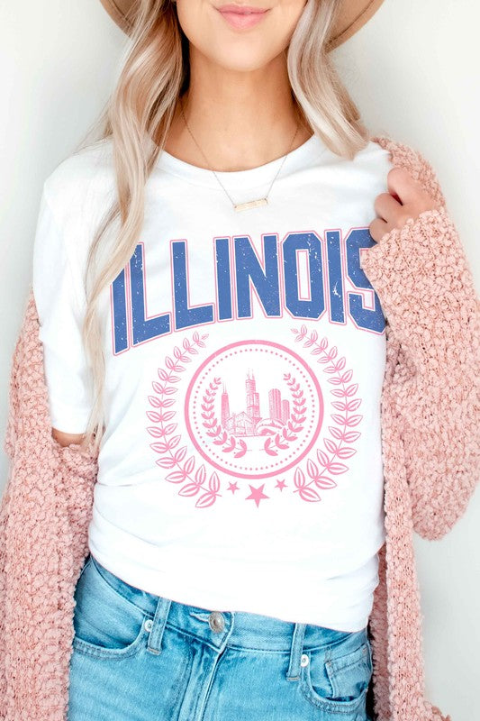 A person is shown wearing an ILLINOIS GRAPHIC TEE, made from 100% cotton, featuring "ILLINOIS" printed in blue above a pink graphic of Chicago's skyline encircled by a decorative laurel wreath; they have wavy hair and are also wearing jeans.