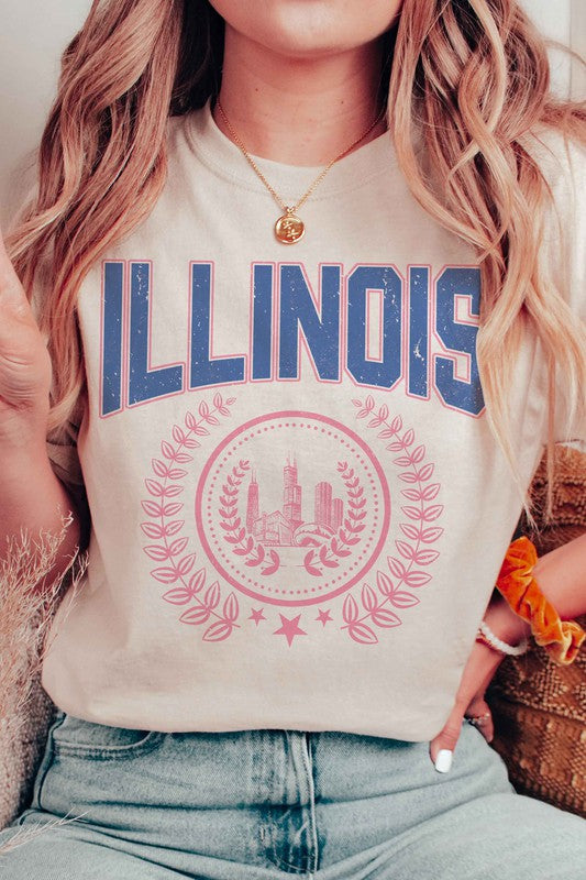 A person is shown wearing an ILLINOIS GRAPHIC TEE, made from 100% cotton, featuring "ILLINOIS" printed in blue above a pink graphic of Chicago's skyline encircled by a decorative laurel wreath; they have wavy hair and are also wearing jeans.