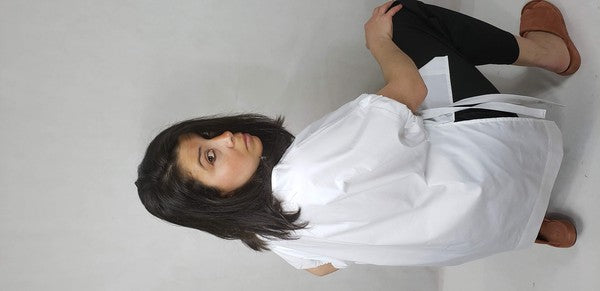 A person kneels on one knee against a neutral background. They wear black pants and a Casual Chic, Crew Neck Top Casual Loose Blouse with ties on the sides. Their right arm is bent upward, and they look off-frame.