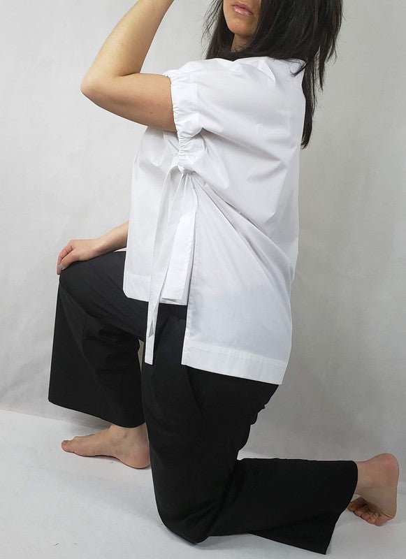 A person kneels on one knee against a neutral background. They wear black pants and a Casual Chic, Crew Neck Top Casual Loose Blouse with ties on the sides. Their right arm is bent upward, and they look off-frame.