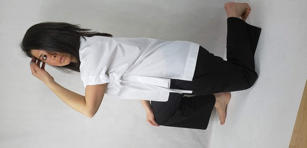 A person kneels on one knee against a neutral background. They wear black pants and a Casual Chic, Crew Neck Top Casual Loose Blouse with ties on the sides. Their right arm is bent upward, and they look off-frame.