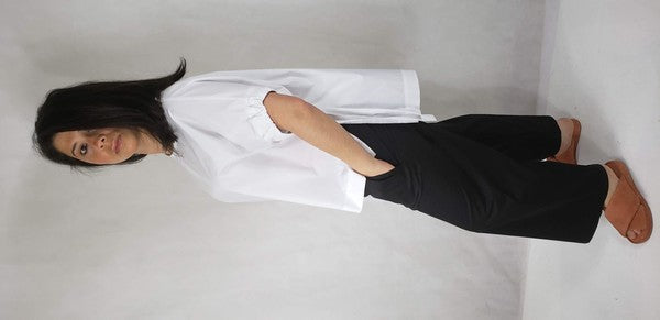 A person kneels on one knee against a neutral background. They wear black pants and a Casual Chic, Crew Neck Top Casual Loose Blouse with ties on the sides. Their right arm is bent upward, and they look off-frame.