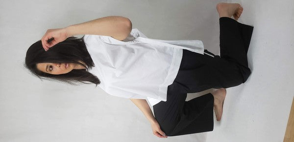 A person kneels on one knee against a neutral background. They wear black pants and a Casual Chic, Crew Neck Top Casual Loose Blouse with ties on the sides. Their right arm is bent upward, and they look off-frame.