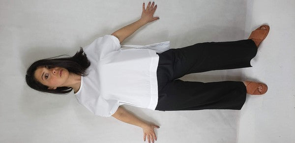 A person kneels on one knee against a neutral background. They wear black pants and a Casual Chic, Crew Neck Top Casual Loose Blouse with ties on the sides. Their right arm is bent upward, and they look off-frame.