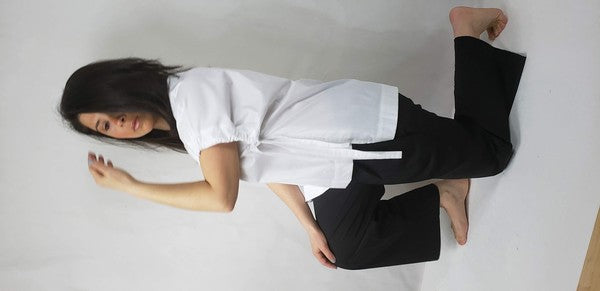 A person kneels on one knee against a neutral background. They wear black pants and a Casual Chic, Crew Neck Top Casual Loose Blouse with ties on the sides. Their right arm is bent upward, and they look off-frame.