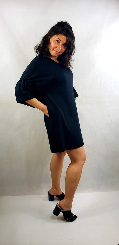 A woman with shoulder-length curly hair stands with her back facing the camera, showcasing a black short sleeve jersey dress with pockets. The loose fit of the dress offers both comfort and style, paired perfectly with her black shoes.