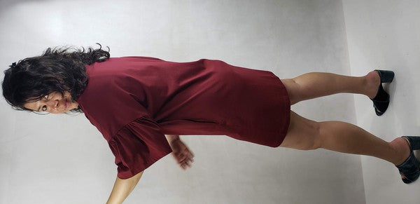 A woman with shoulder-length curly hair stands with her back facing the camera, showcasing a black short sleeve jersey dress with pockets. The loose fit of the dress offers both comfort and style, paired perfectly with her black shoes.