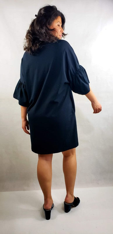 A woman with shoulder-length curly hair stands with her back facing the camera, showcasing a black short sleeve jersey dress with pockets. The loose fit of the dress offers both comfort and style, paired perfectly with her black shoes.