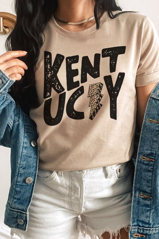 Person standing, wearing the KENTUCKY LEOPARD LIGHTNING GRAPHIC TEE, a denim jacket draped over their shoulders, and white shorts.