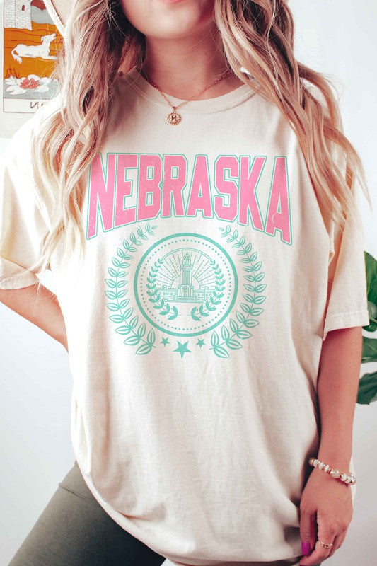 Person wearing the NEBRASKA GRAPHIC TEE in white, which features "Nebraska" in pink letters and a design beneath it, paired with white shorts. Made from 100% cotton, this tee ensures comfort and quality, making it perfect for plus-size fashion enthusiasts.