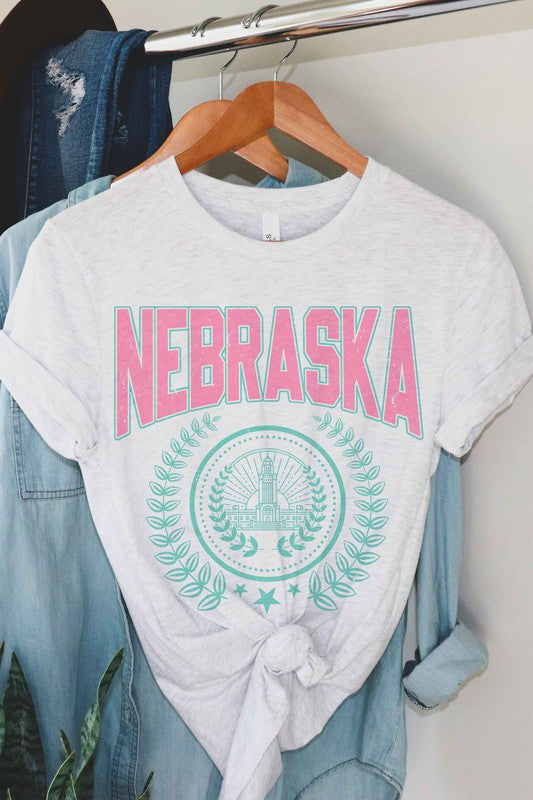 Person wearing the NEBRASKA GRAPHIC TEE in white, which features "Nebraska" in pink letters and a design beneath it, paired with white shorts. Made from 100% cotton, this tee ensures comfort and quality, making it perfect for plus-size fashion enthusiasts.