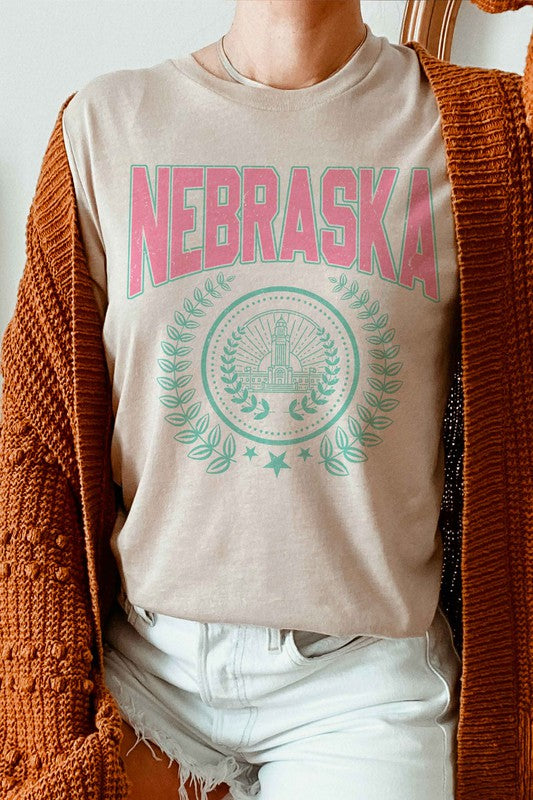 Person wearing the NEBRASKA GRAPHIC TEE in white, which features "Nebraska" in pink letters and a design beneath it, paired with white shorts. Made from 100% cotton, this tee ensures comfort and quality, making it perfect for plus-size fashion enthusiasts.