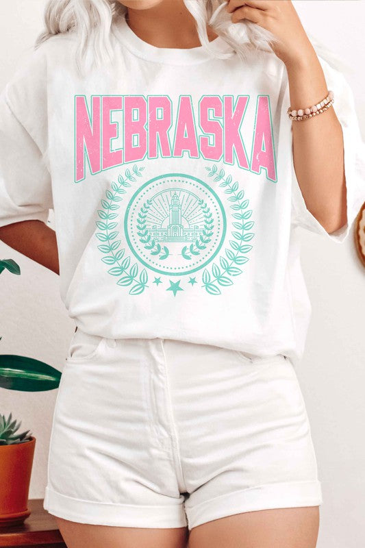 Person wearing the NEBRASKA GRAPHIC TEE in white, which features "Nebraska" in pink letters and a design beneath it, paired with white shorts. Made from 100% cotton, this tee ensures comfort and quality, making it perfect for plus-size fashion enthusiasts.