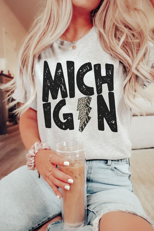 Someone sporting a MICHIGAN LEOPARD LIGHTNING GRAPHIC TEE and holding a drink, with the "I" in Michigan uniquely designed as a lightning bolt.