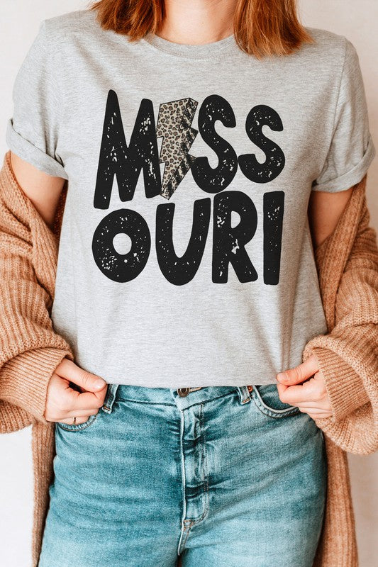 A person wearing a beige MISSOURI LEOPARD LIGHTNING GRAPHIC TEE in unisex sizing, sitting on a woven chair, rocking blue jeans, a bracelet, and a necklace with a circular pendant.