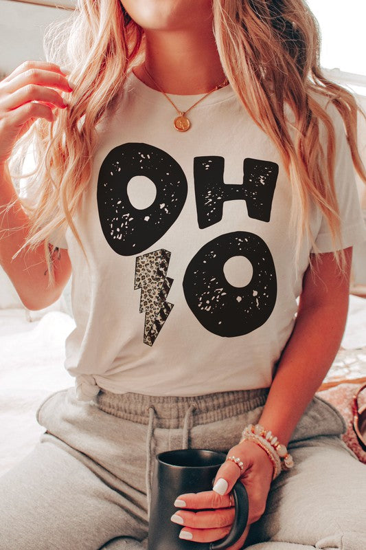 A person is sporting an OHIO LEOPARD LIGHTNING GRAPHIC TEE, which showcases large distressed black letters spelling "OH" and "HO" divided by a leopard print lightning bolt. They have a denim jacket casually draped over their shoulders and are paired with light gray pants. This unisex t-shirt is designed to be stylishly inclusive, available in plus sizes.