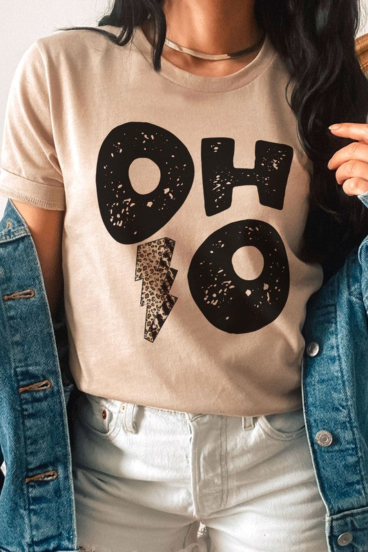 A person is sporting an OHIO LEOPARD LIGHTNING GRAPHIC TEE, which showcases large distressed black letters spelling "OH" and "HO" divided by a leopard print lightning bolt. They have a denim jacket casually draped over their shoulders and are paired with light gray pants. This unisex t-shirt is designed to be stylishly inclusive, available in plus sizes.