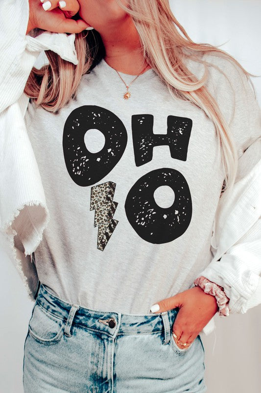 A person is sporting an OHIO LEOPARD LIGHTNING GRAPHIC TEE, which showcases large distressed black letters spelling "OH" and "HO" divided by a leopard print lightning bolt. They have a denim jacket casually draped over their shoulders and are paired with light gray pants. This unisex t-shirt is designed to be stylishly inclusive, available in plus sizes.