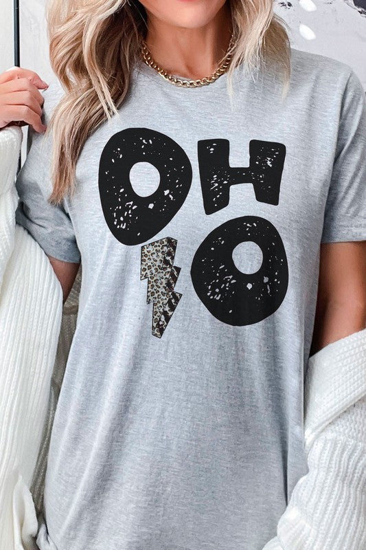 A person is sporting an OHIO LEOPARD LIGHTNING GRAPHIC TEE, which showcases large distressed black letters spelling "OH" and "HO" divided by a leopard print lightning bolt. They have a denim jacket casually draped over their shoulders and are paired with light gray pants. This unisex t-shirt is designed to be stylishly inclusive, available in plus sizes.