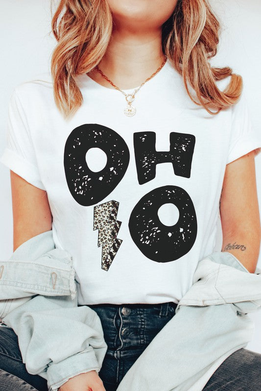 A person is sporting an OHIO LEOPARD LIGHTNING GRAPHIC TEE, which showcases large distressed black letters spelling "OH" and "HO" divided by a leopard print lightning bolt. They have a denim jacket casually draped over their shoulders and are paired with light gray pants. This unisex t-shirt is designed to be stylishly inclusive, available in plus sizes.