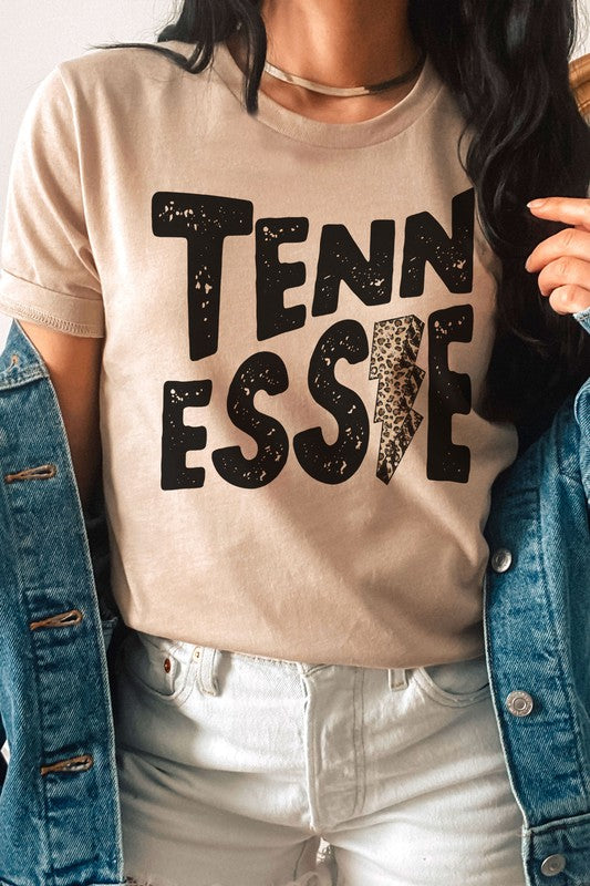 Wearing a gray TENNESSEE Leopard Lightning Graphic Tee with the word "TENNESSEE" prominently displayed in bold black letters and a lightning bolt design within the letter "E," this stylish unisex t-shirt provides an inclusive fashion choice for everyone.