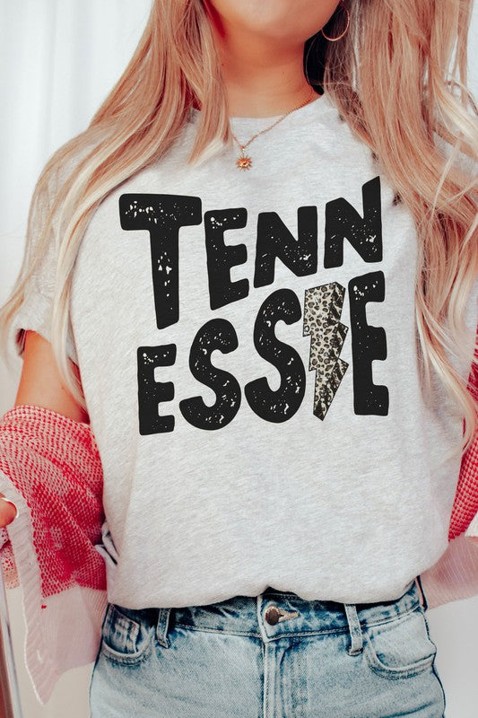 Wearing a gray TENNESSEE Leopard Lightning Graphic Tee with the word "TENNESSEE" prominently displayed in bold black letters and a lightning bolt design within the letter "E," this stylish unisex t-shirt provides an inclusive fashion choice for everyone.