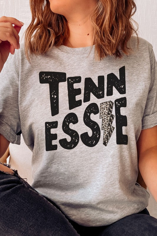 Wearing a gray TENNESSEE Leopard Lightning Graphic Tee with the word "TENNESSEE" prominently displayed in bold black letters and a lightning bolt design within the letter "E," this stylish unisex t-shirt provides an inclusive fashion choice for everyone.