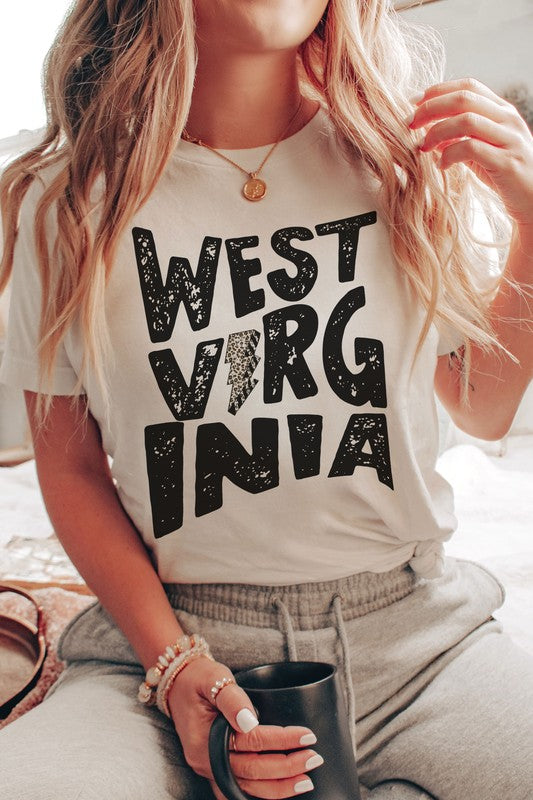 Person with long hair wearing a white "West Virginia Leopard Lightning Graphic Tee" and jeans stands indoors, looking like they just came from a thrilling football game.