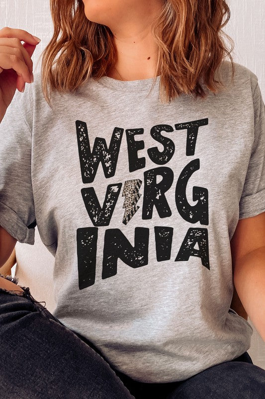 Person with long hair wearing a white "West Virginia Leopard Lightning Graphic Tee" and jeans stands indoors, looking like they just came from a thrilling football game.