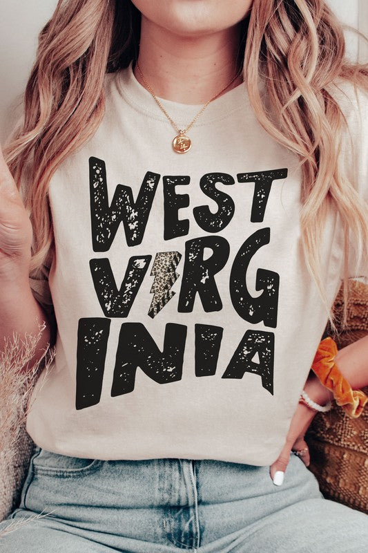 Person with long hair wearing a white "West Virginia Leopard Lightning Graphic Tee" and jeans stands indoors, looking like they just came from a thrilling football game.