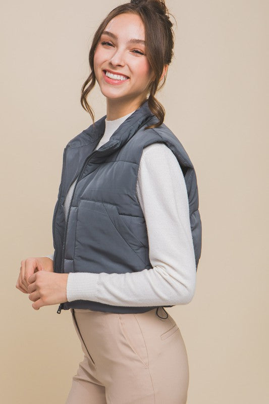 A young woman wearing the Puffer Vest With Pockets in beige, featuring a quilted design, over a white long-sleeve ribbed top and light blue jeans stands against a plain background.