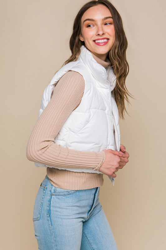 A young woman wearing the Puffer Vest With Pockets in beige, featuring a quilted design, over a white long-sleeve ribbed top and light blue jeans stands against a plain background.