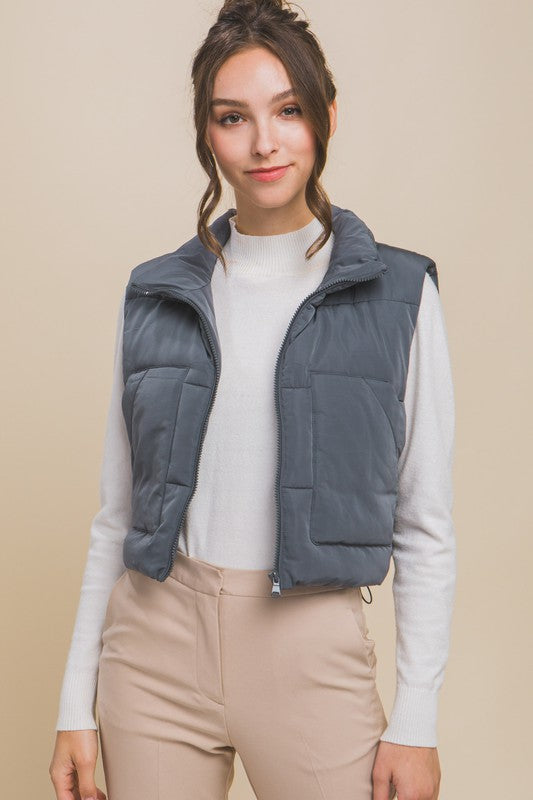 A young woman wearing the Puffer Vest With Pockets in beige, featuring a quilted design, over a white long-sleeve ribbed top and light blue jeans stands against a plain background.