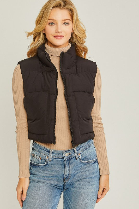 A young woman wearing the Puffer Vest With Pockets in beige, featuring a quilted design, over a white long-sleeve ribbed top and light blue jeans stands against a plain background.