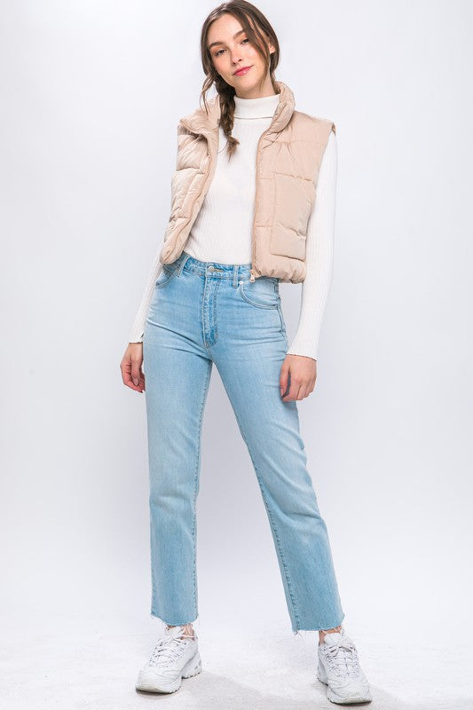 A young woman wearing the Puffer Vest With Pockets in beige, featuring a quilted design, over a white long-sleeve ribbed top and light blue jeans stands against a plain background.