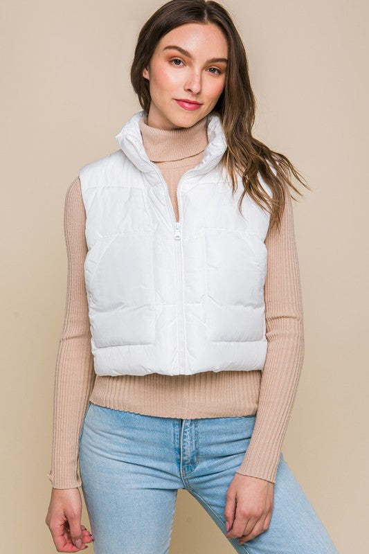 A young woman wearing the Puffer Vest With Pockets in beige, featuring a quilted design, over a white long-sleeve ribbed top and light blue jeans stands against a plain background.