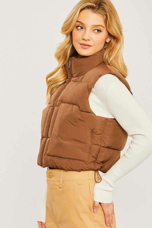 A young woman wearing the Puffer Vest With Pockets in beige, featuring a quilted design, over a white long-sleeve ribbed top and light blue jeans stands against a plain background.