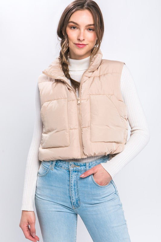 A young woman wearing the Puffer Vest With Pockets in beige, featuring a quilted design, over a white long-sleeve ribbed top and light blue jeans stands against a plain background.