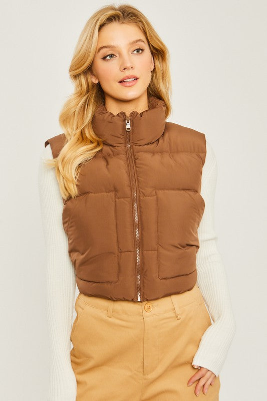 A young woman wearing the Puffer Vest With Pockets in beige, featuring a quilted design, over a white long-sleeve ribbed top and light blue jeans stands against a plain background.