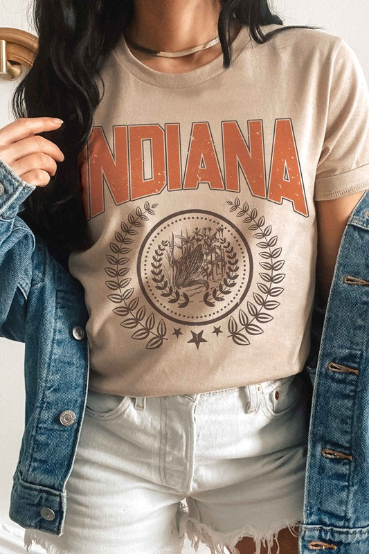 Person wearing a beige INDIANA GRAPHIC TEE made from 100% cotton, with a detailed graphic below it, white shorts, and a denim jacket draped over their shoulders.