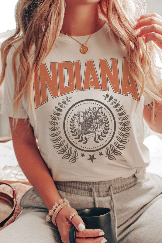 Person wearing a beige INDIANA GRAPHIC TEE made from 100% cotton, with a detailed graphic below it, white shorts, and a denim jacket draped over their shoulders.