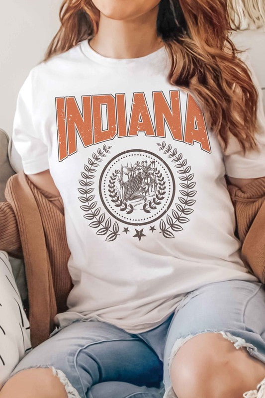 Person wearing a beige INDIANA GRAPHIC TEE made from 100% cotton, with a detailed graphic below it, white shorts, and a denim jacket draped over their shoulders.