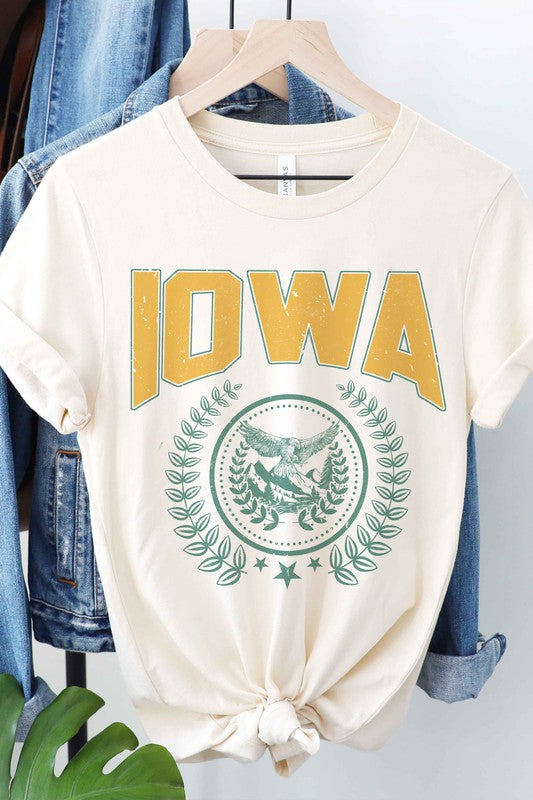 The IOWA GRAPHIC TEE in white showcases "IOWA" in bold yellow letters at the top, accompanied by an eagle graphic surrounded by greenery and stars below. This unisex t-shirt, also available in plus sizes, is displayed on a white surface beside denim shorts.