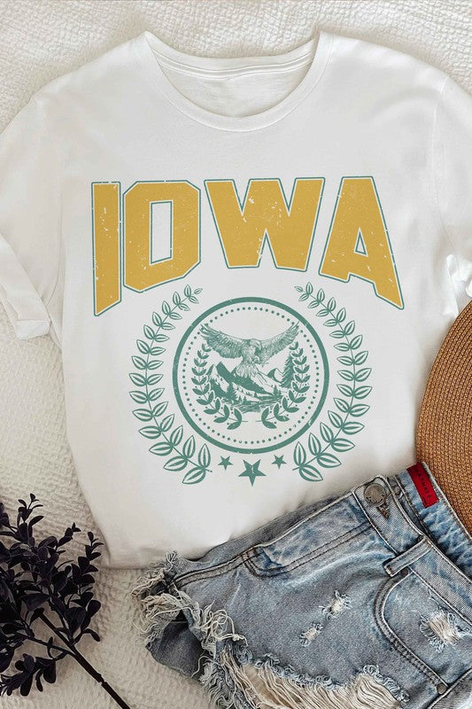 The IOWA GRAPHIC TEE in white showcases "IOWA" in bold yellow letters at the top, accompanied by an eagle graphic surrounded by greenery and stars below. This unisex t-shirt, also available in plus sizes, is displayed on a white surface beside denim shorts.