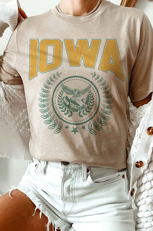 The IOWA GRAPHIC TEE in white showcases "IOWA" in bold yellow letters at the top, accompanied by an eagle graphic surrounded by greenery and stars below. This unisex t-shirt, also available in plus sizes, is displayed on a white surface beside denim shorts.