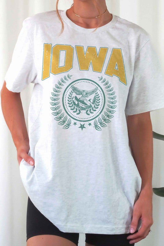 The IOWA GRAPHIC TEE in white showcases "IOWA" in bold yellow letters at the top, accompanied by an eagle graphic surrounded by greenery and stars below. This unisex t-shirt, also available in plus sizes, is displayed on a white surface beside denim shorts.