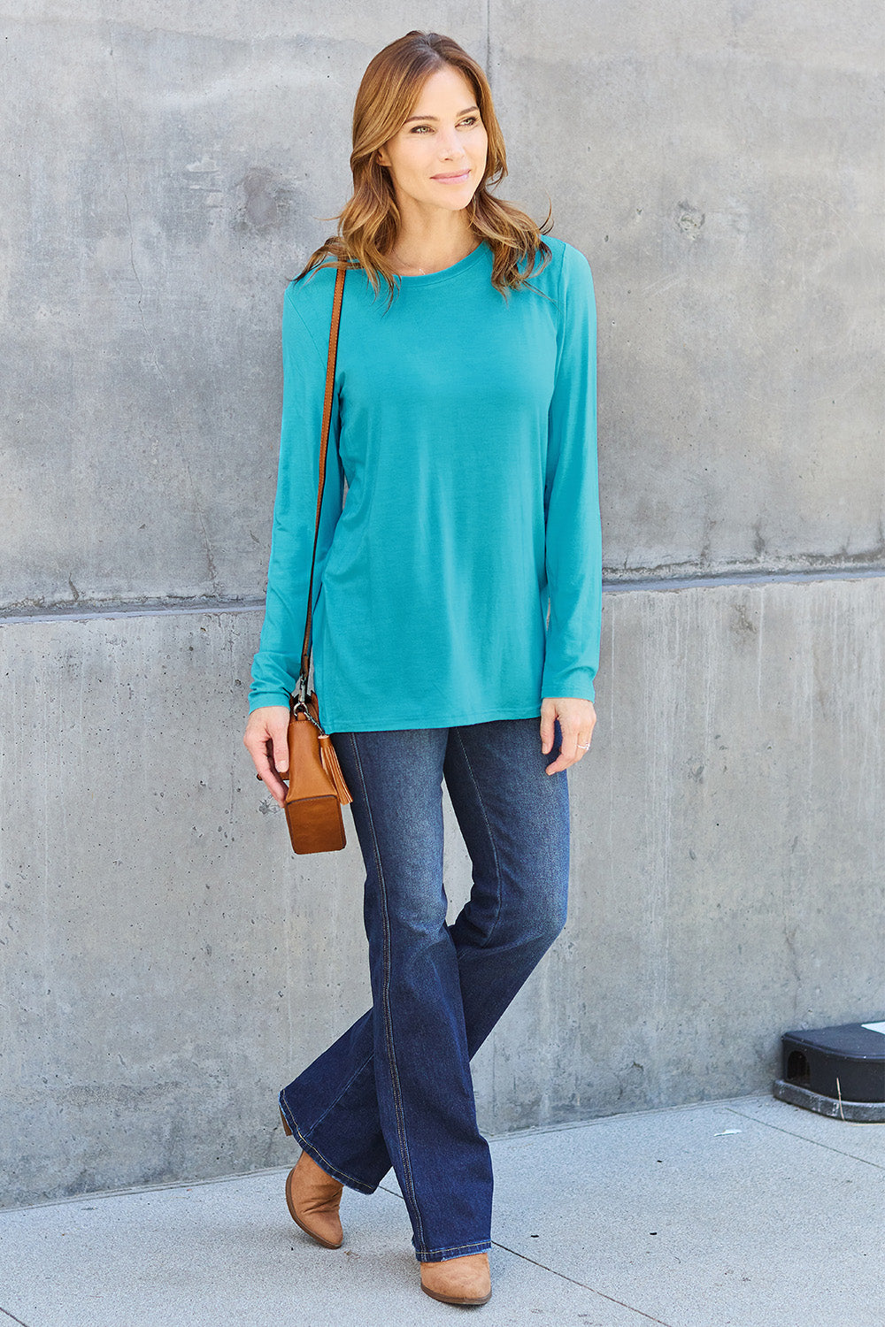 A woman with shoulder-length hair wearing the Basic Bae Full Size Round Neck Long Sleeve Top and blue jeans stands in front of a concrete wall, holding a beige handbag over her shoulder. Her outfit exemplifies basic style, with the top made from 100% polyester and recommended for machine wash cold.