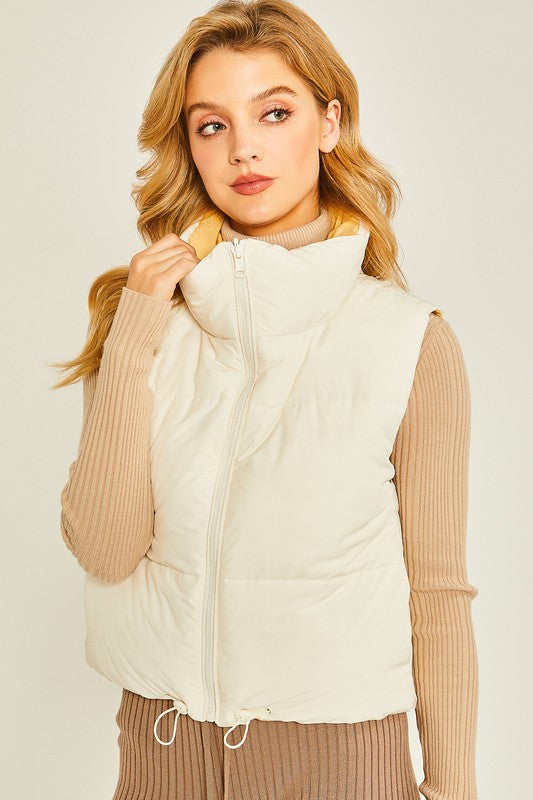 A person wearing a beige turtleneck sweater and a Woven Solid Reversible Vest with a versatile style stands against a plain background, holding the collar of the vest with one hand.