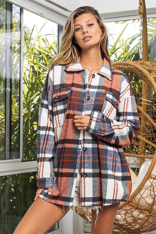 A woman in a Textured Shirt With Big Checkered Point leans casually against a wall, smiling while looking to the side. There are plants and a window in the background.