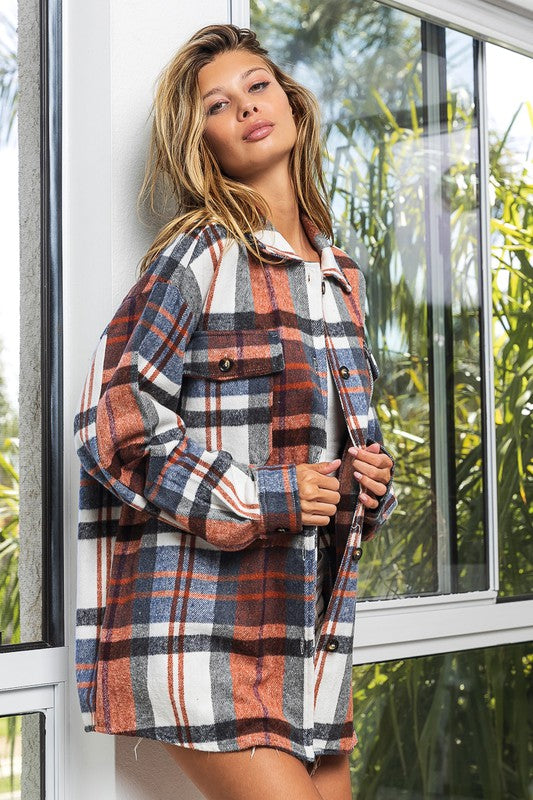 A woman in a Textured Shirt With Big Checkered Point leans casually against a wall, smiling while looking to the side. There are plants and a window in the background.